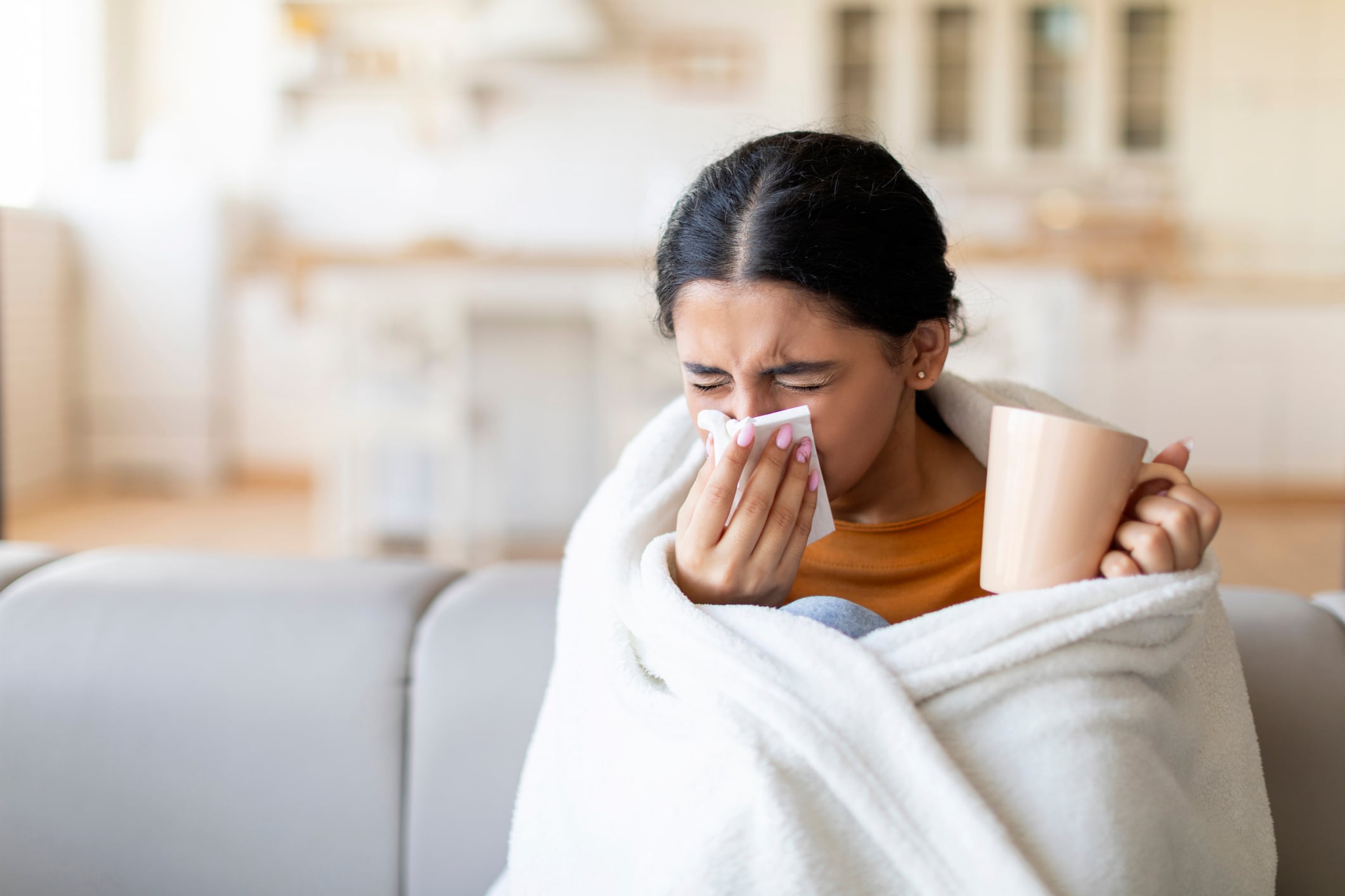Woman wrapped in blanket on the sofa, holding a mug and blowing her nose