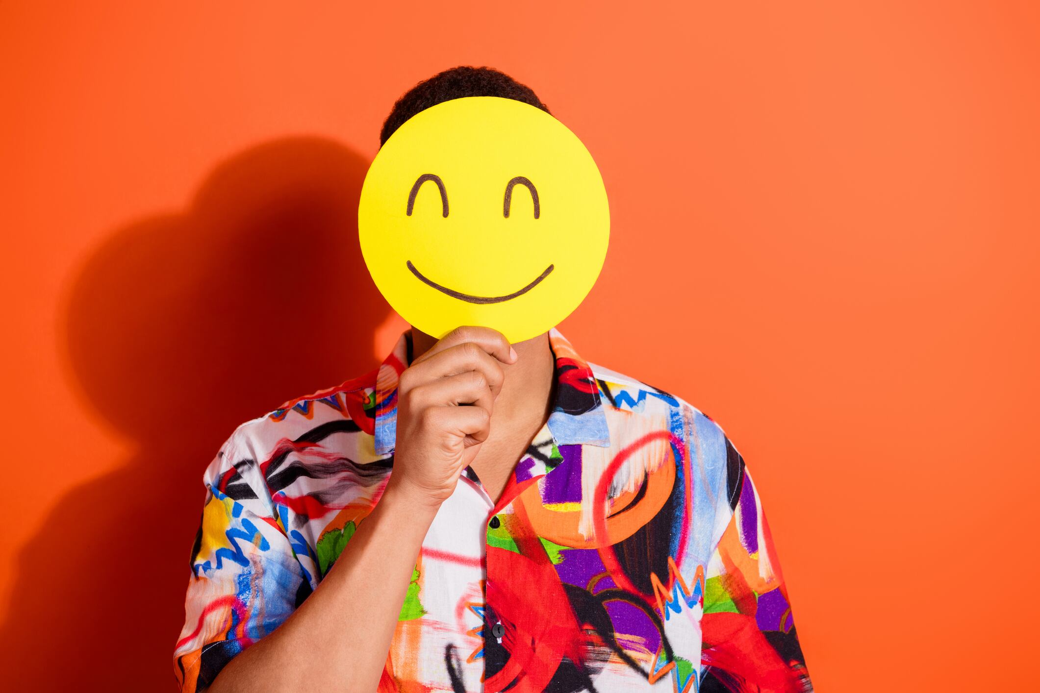 A young man holds a yellow smiley face, partially hiding his face against a vibrant orange background. He wears a stylish, colorful shirt, creating a cheerful and trendy image.