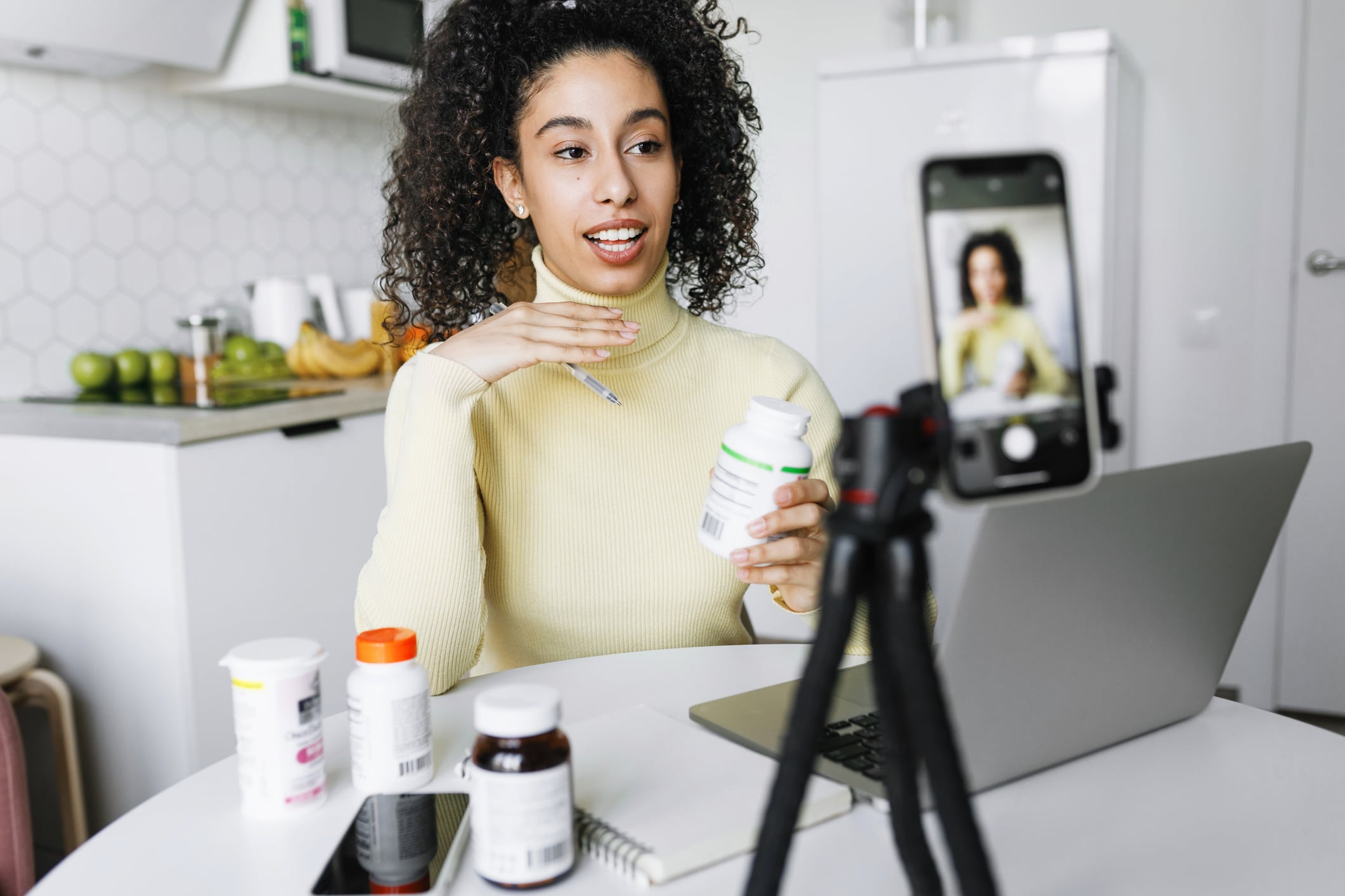 Female blogger nutritionist recording video tutorial with advice and tips on immune boosting techniques on smartphone camera recommending food supplements, holding bottle in hand