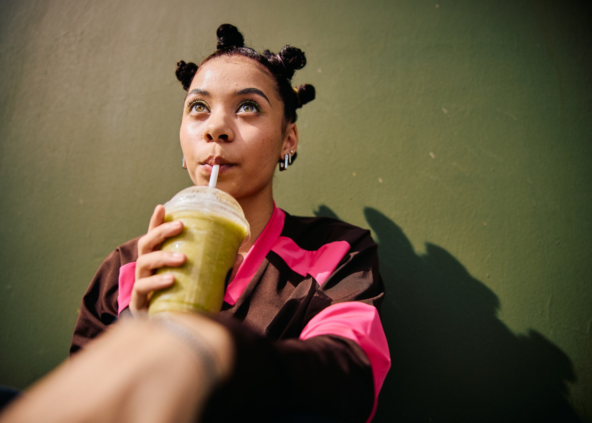 Closeup of a teenage girl with bantu knots holding a takeaway cup and drinking a smoothie while looking up against a plain green wall in bright sunlight.