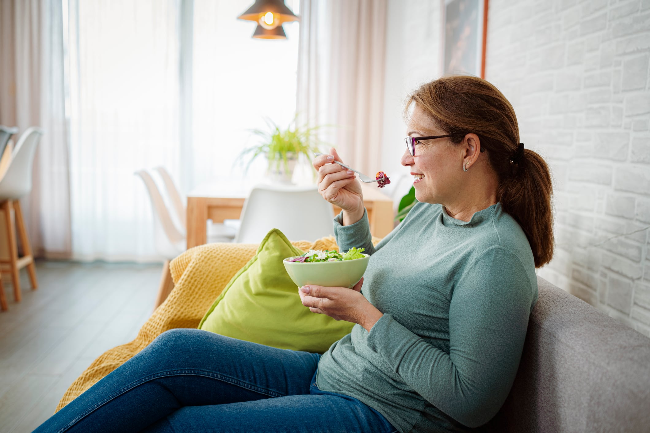 Side view of mature hispanic woman eating healthy vegetables salad sitting on sofa