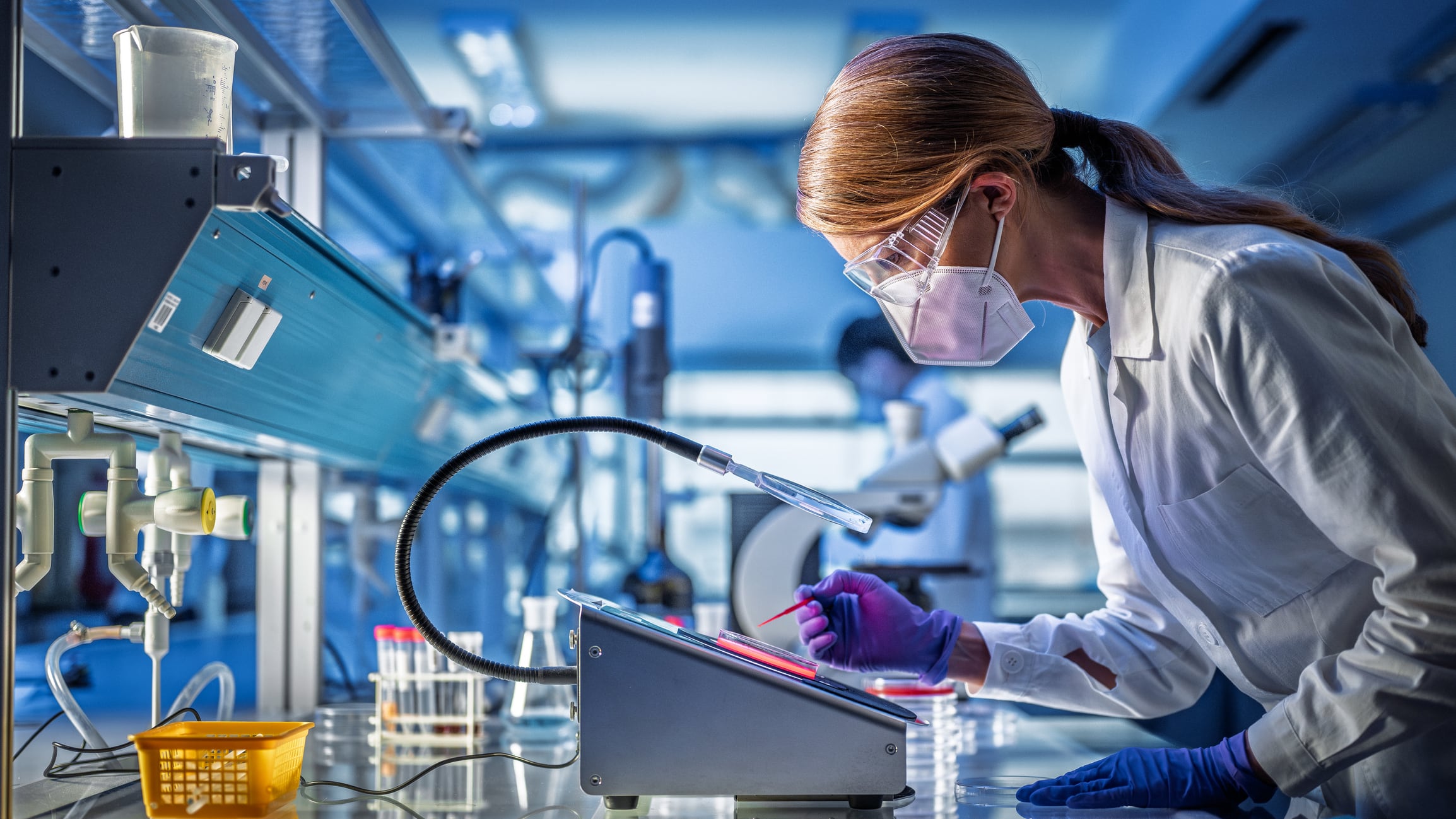 Mature Caucasian female professor examining sample at laboratory bench, wearing protective goggles, gloves and lab coat. Sterile workspace with scientific equipment and containers visible. Professional research environment with controlled lighting.