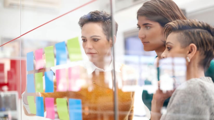 Three women of different ages looking at different colored post it notes on a board to depict menopause market segmentation
