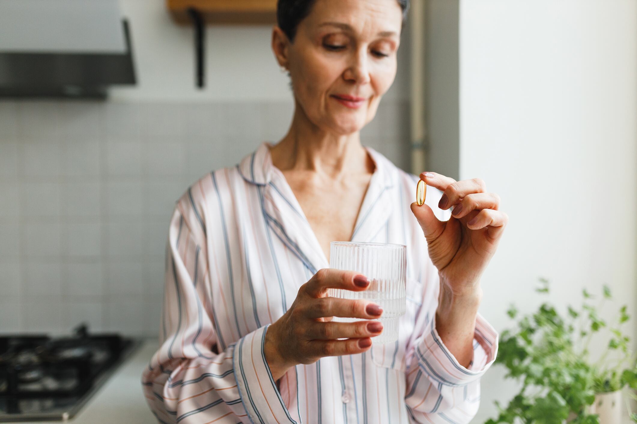 Daily routine of middle-aged healthy and good-looking female for collagen and antioxidant support based on taking dietary supplements, woman holding gel capsule of fish oil and glass of water