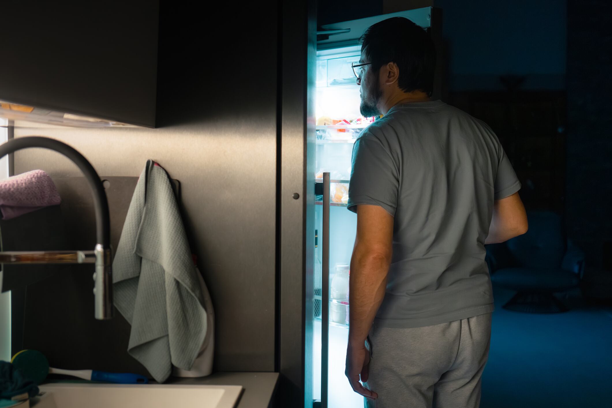 Man standing in a dark kitchen, the refrigerator light illuminating his face as he looks for a snack