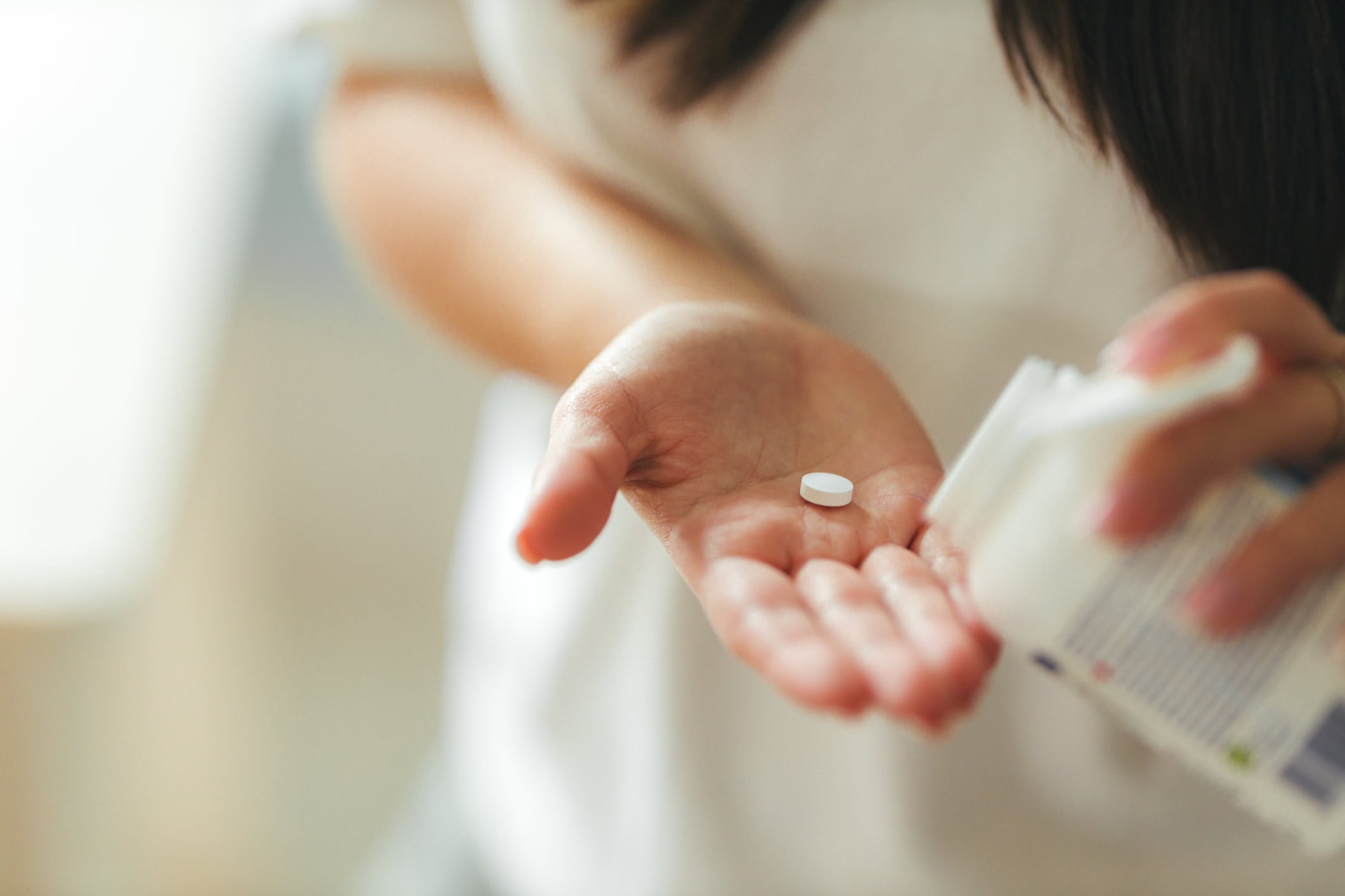 Close-up of young adult woman at home during daytime pouring a vitamin tablet from a supplement bottle into her hand. Casual clothing, shallow depth of field.