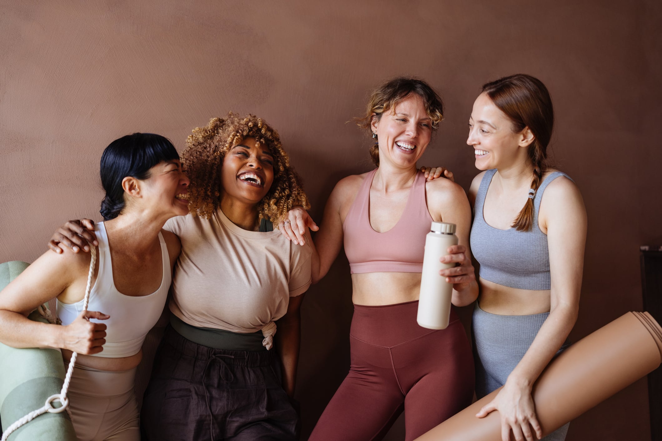 A group of four women in athletic wear share a joyful moment after a workout, showcasing friendship, fitness, and teamwork in a warm studio setting.