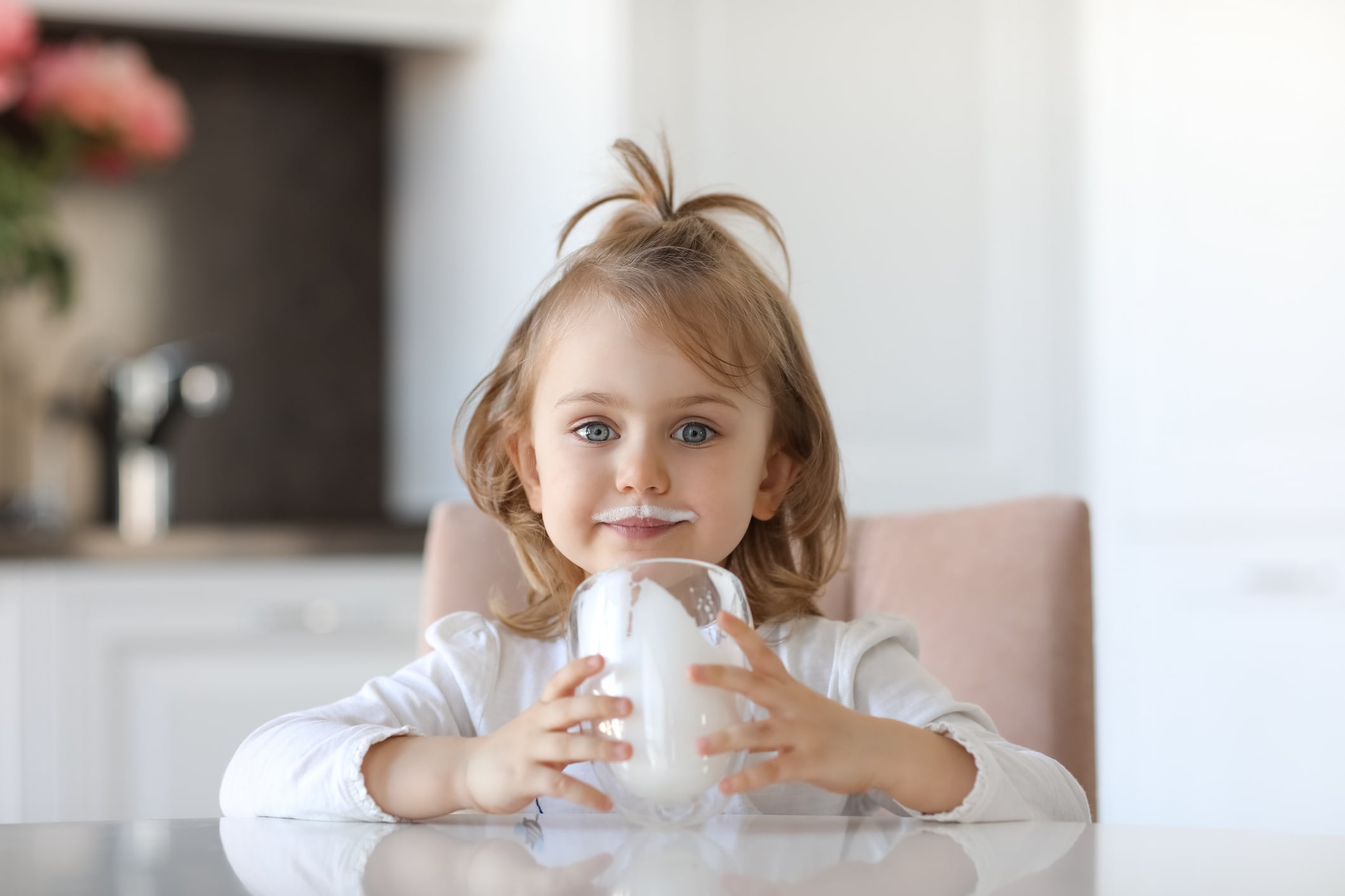 Girl with traces of milk on the lips is holding a glass of milk siting at a white table in a white kitchen. Milk for good health