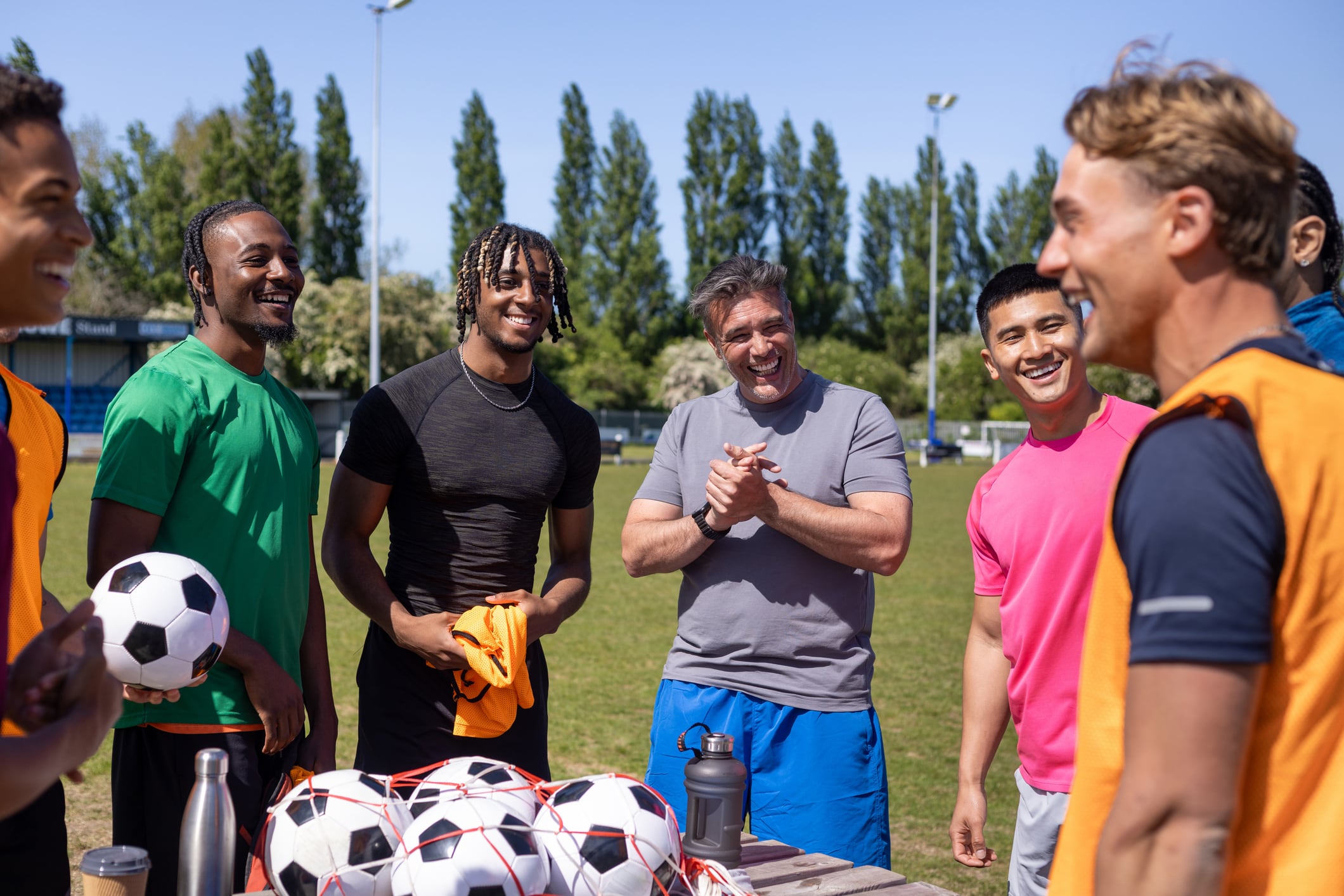 A wide view of a mature football coach talking to a diverse group of young male amateur footballers, they are standing around a table near the football pitch at football training in the North East of England. The coach is giving a pep talk before the team starts football practice. They are wearing sports clothing.