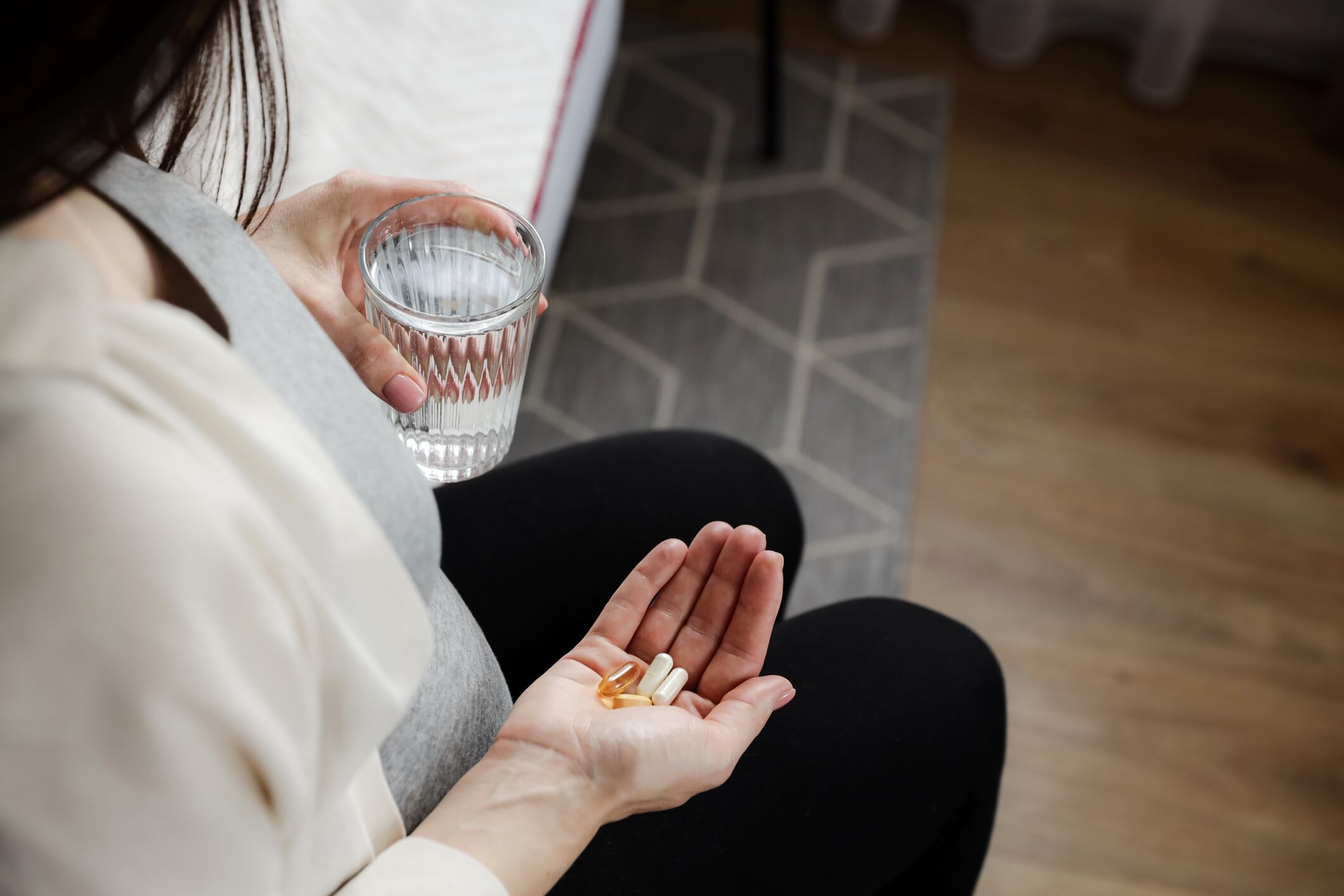 Close up of pregnant woman holding glass of water and heap of pills in hand, taking vitamins D, E, A, calcium, dietary supplements, omega 3 in fish oil capsules at home. Pregnancy, prenatal health