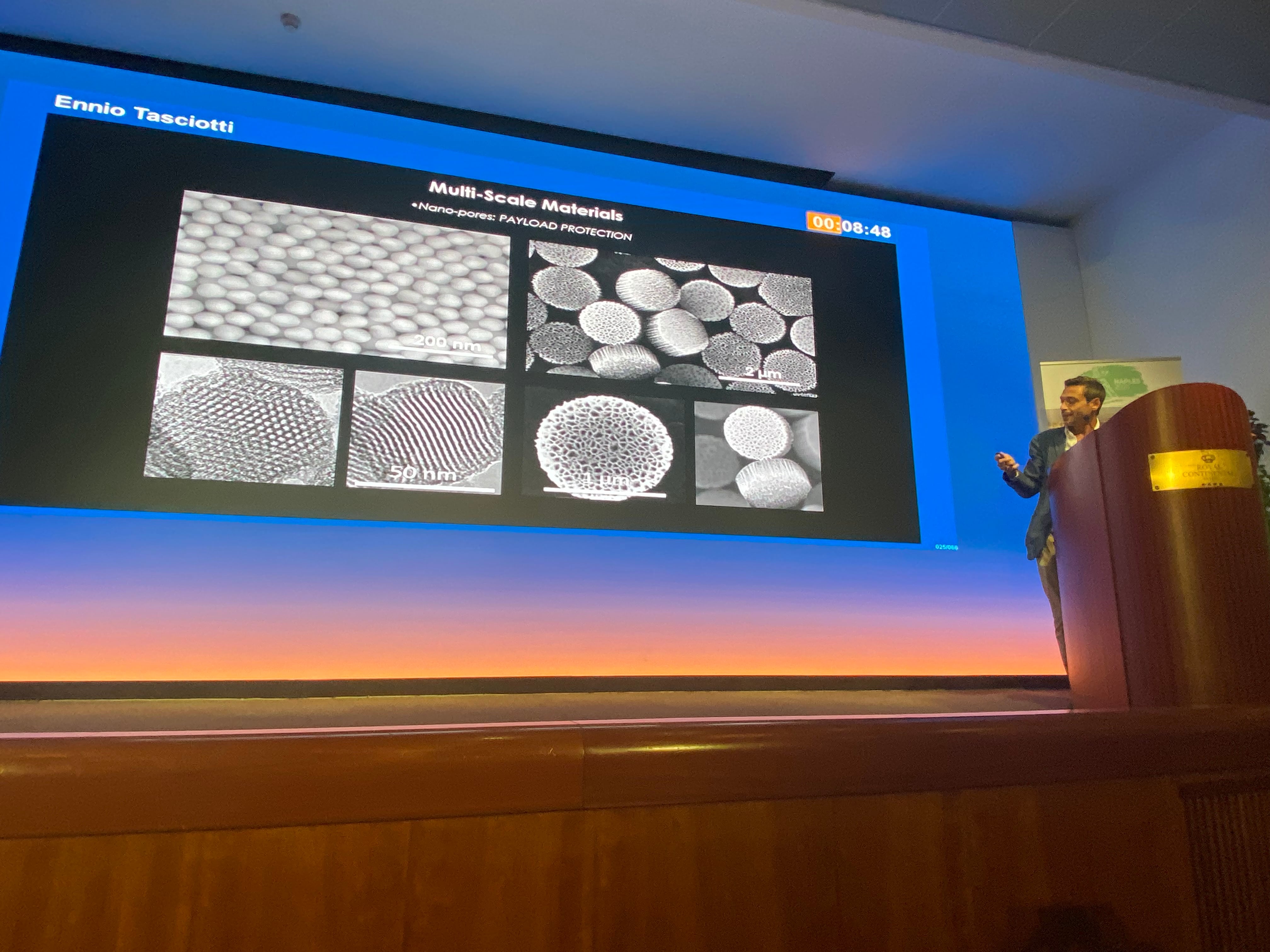 Prof. Ennio Tasciotti on stage at GA Naples presenting a slide on multi-scale materials with nano-pores for payload protection.