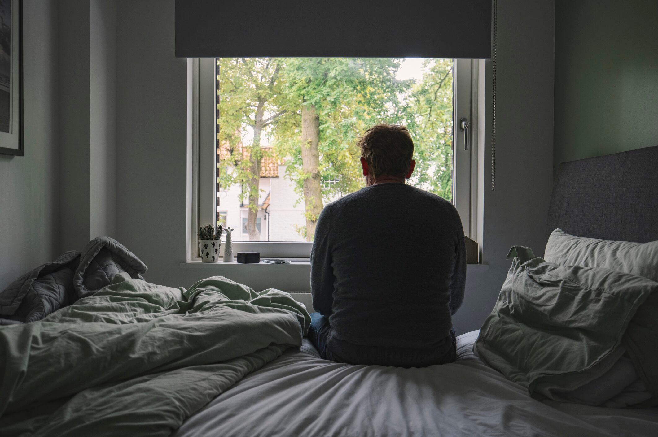 Rear view of depressed man sitting on bed and looking out the window