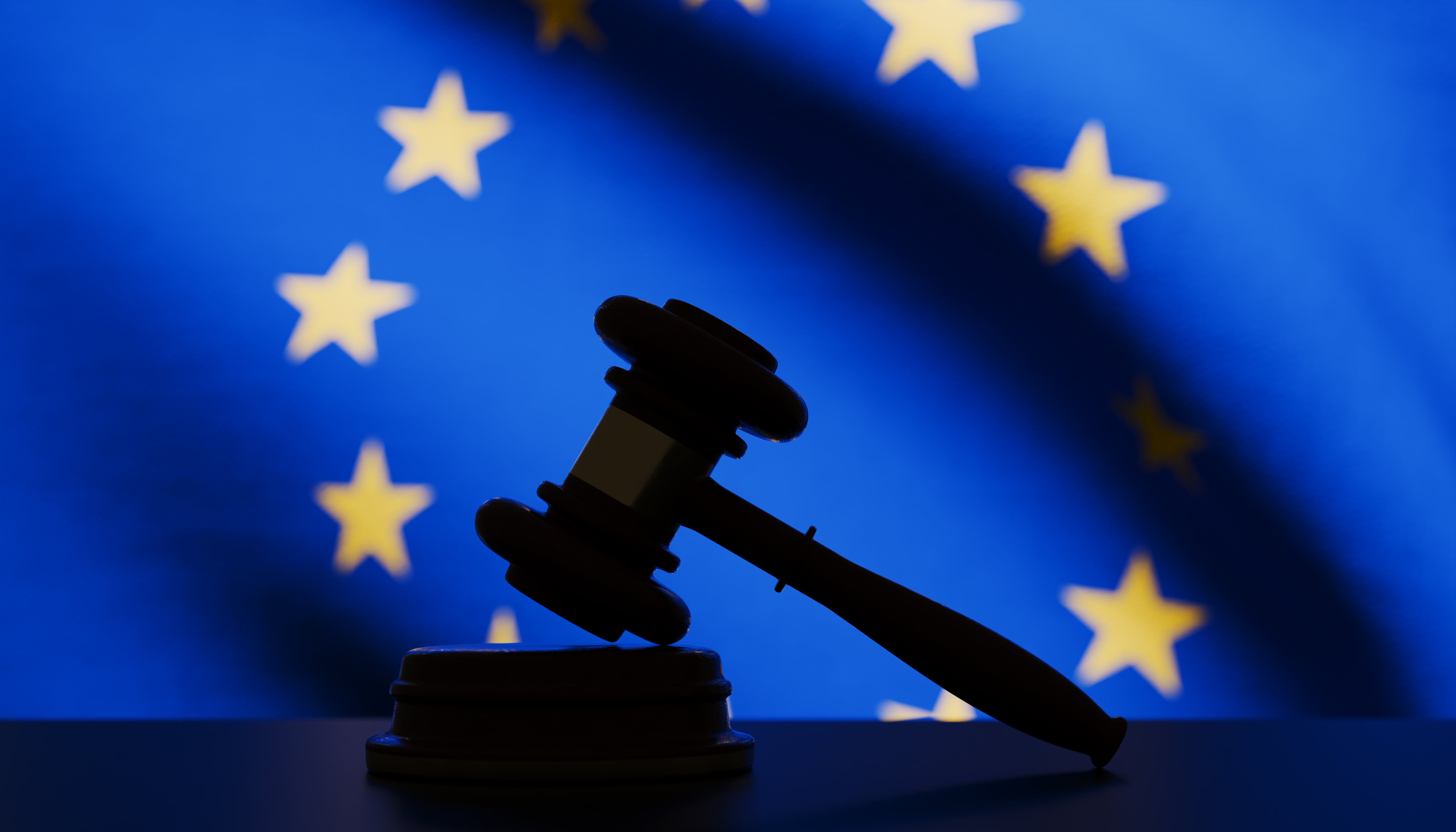 A gavel is shown in silhouette against the backdrop of the European Union flag, its dark outline contrasting with the blue field and yellow stars. The image symbolizes justice, law, and authority across the EU member states, emphasizing themes of unity, governance, and the rule of law within the European Union. The combination of the gavel and the EU flag highlights the role of legal frameworks and cooperation in maintaining order and justice across Europe.