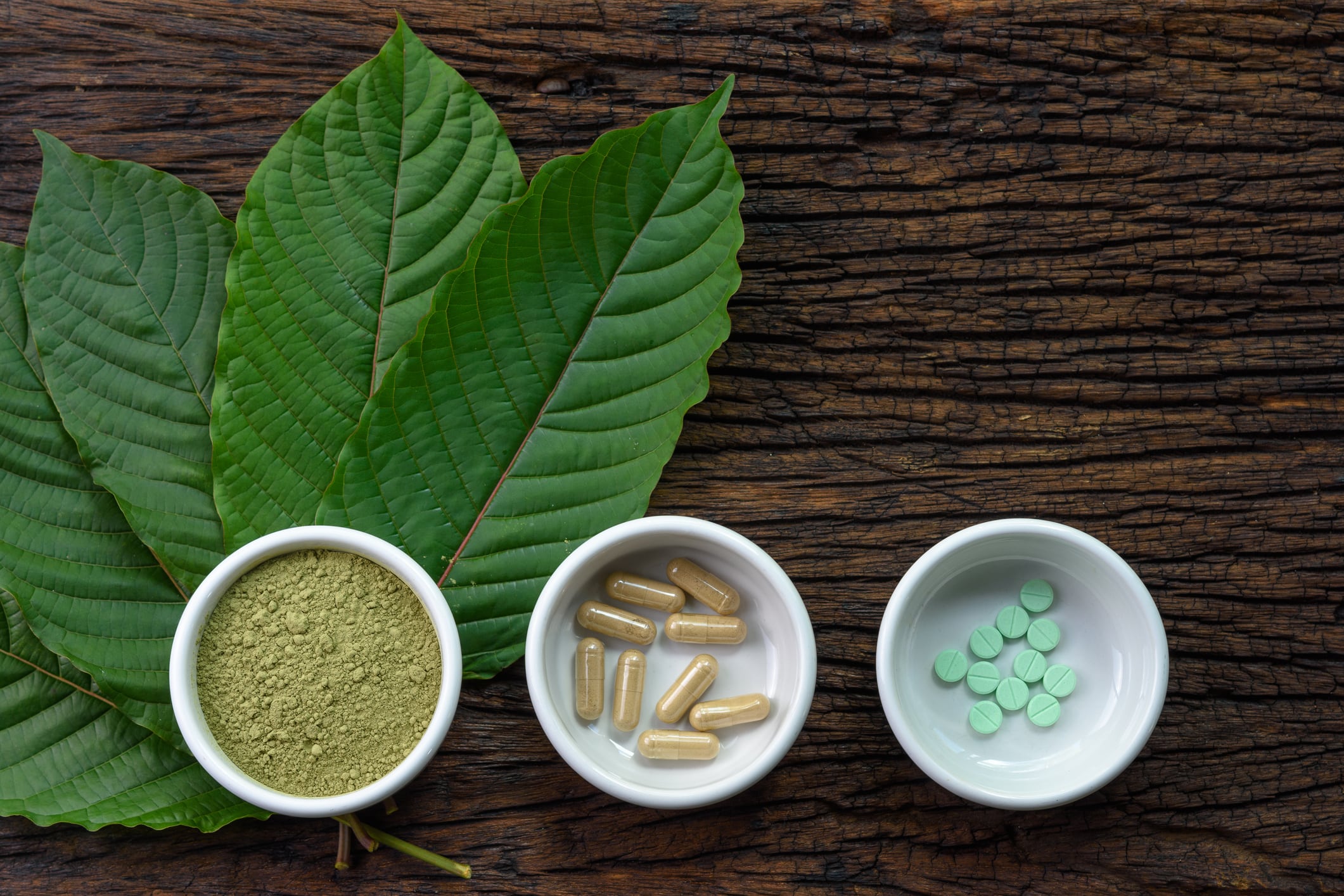 Mitragyna speciosa (kratom) leaves with medicine products in powder, capsules and tablet in white ceramic bowl with wooden texture on background