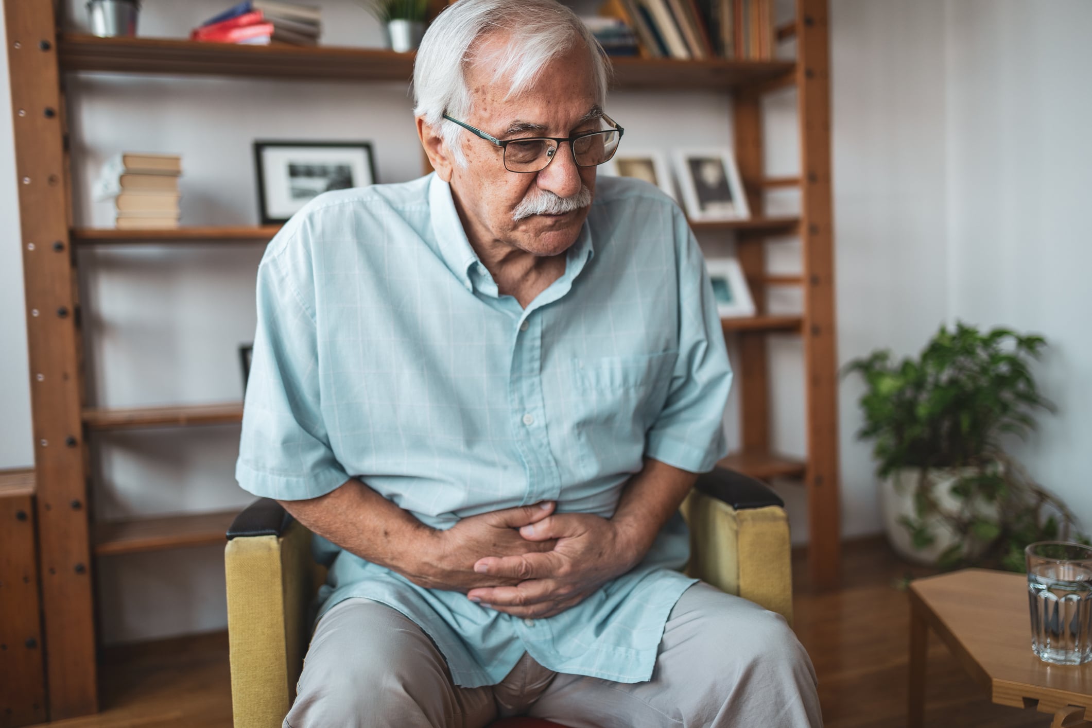 Senior man sitting in his armchair holding hand on his stomach because he is feeling pain