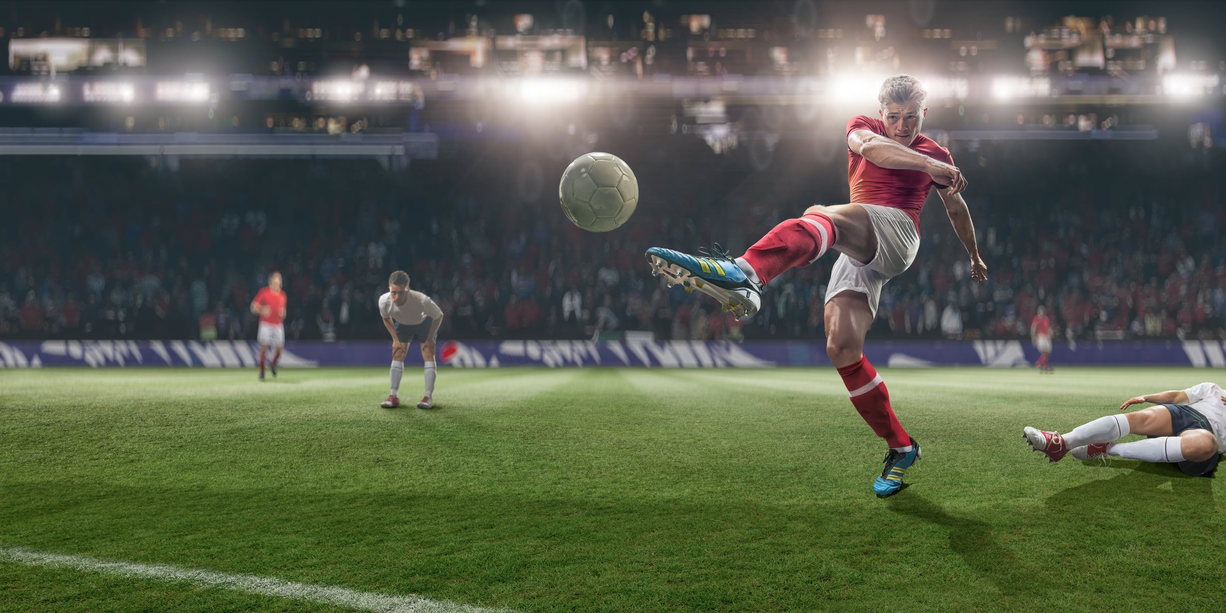 A mid action image of soccer player kicking football, following a sliding tackle challenge from a rival player. The action takes place on a soccer pitch on a generic outdoor floodlit football stadium full of spectators. All players are wearing unbranded generic kit.