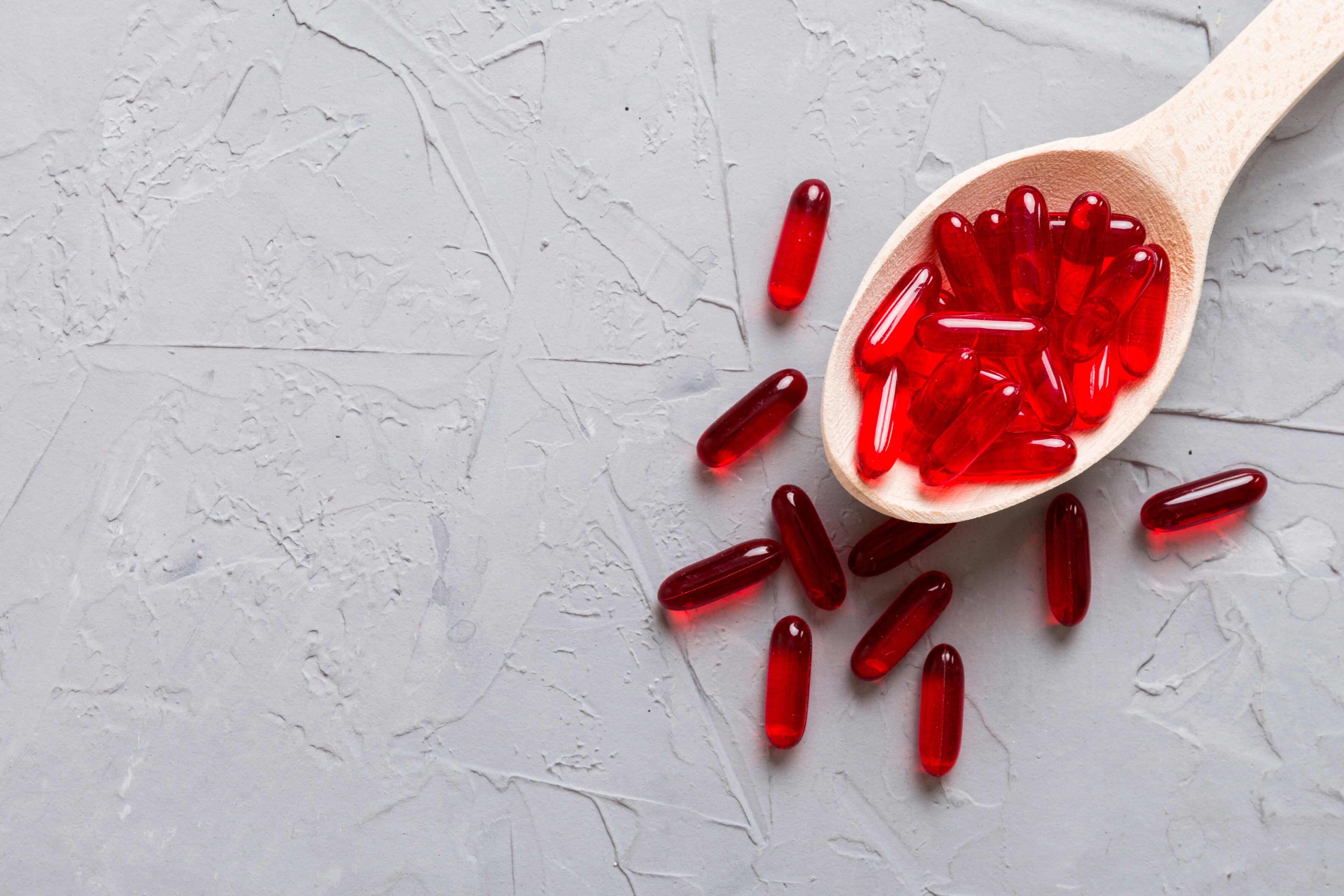 Vitamin capsules in a spoon on a colored background. Pills served as a healthy meal. Red soft gel vitamin supplement capsules on spoon.