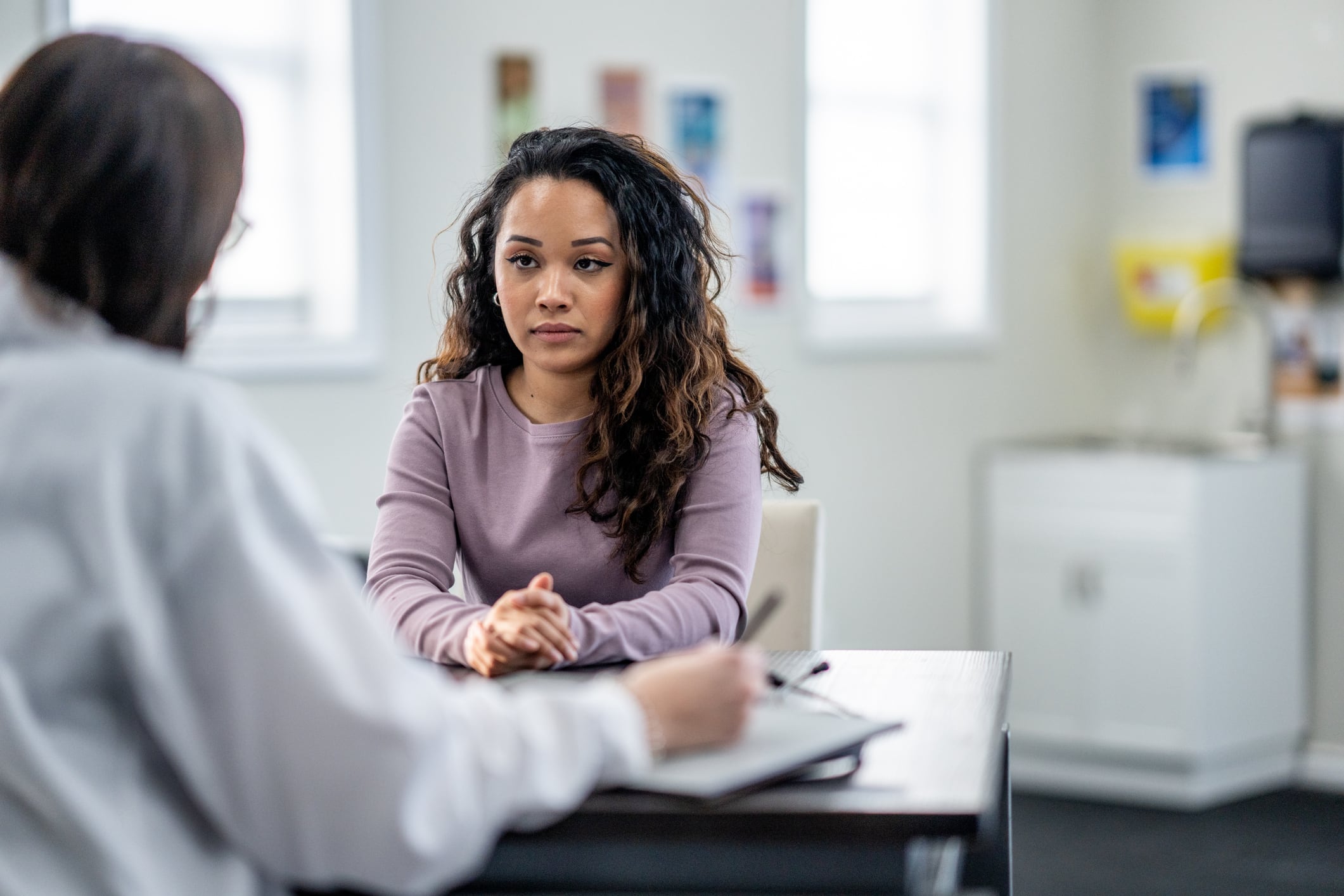 A young woman of mixed race, sits with her female doctor as they discuss her mental health.  The patient is dressed casually and appears depressed as she looks down low with a forlorn expression on her face.