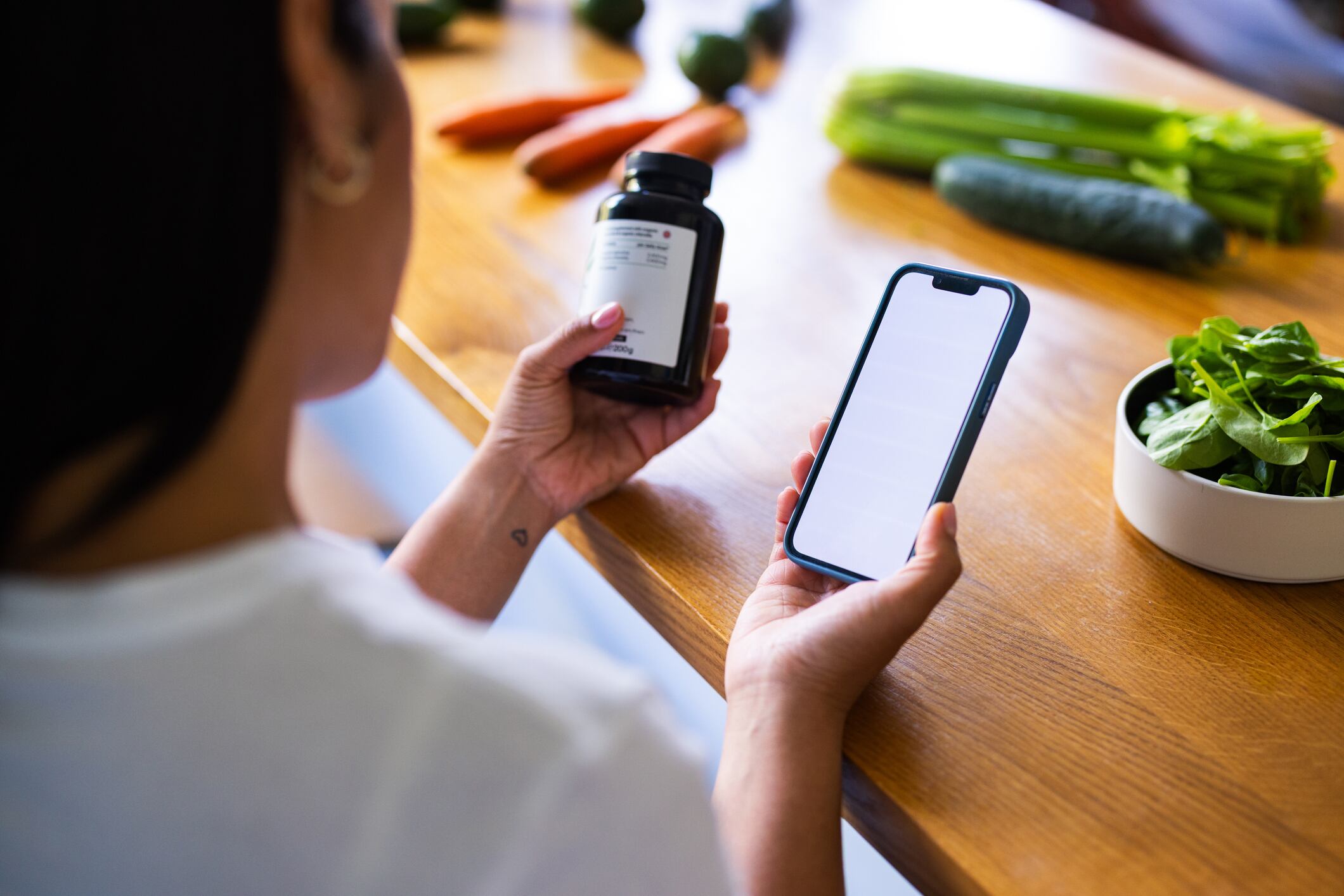 A person sits at a wooden kitchen counter, looking at a smartphone while holding a supplement bottle. Fresh vegetables in the background create a healthy, home lifestyle scene.