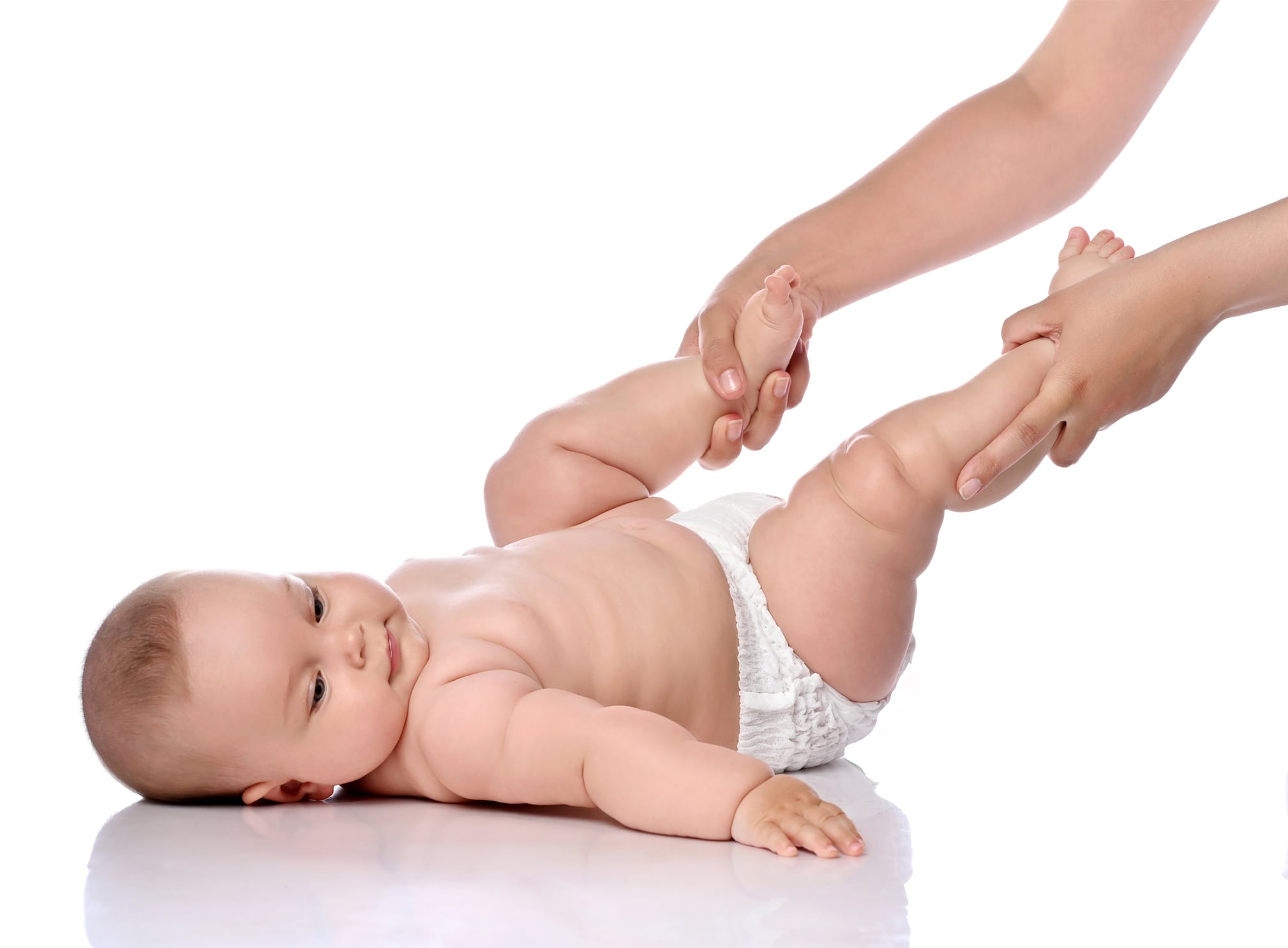 Calm, smiling infant child baby girl kid in diaper is lying on her back doing exercises to strengthen legs and back muscles with her mother, coach holding her feet up isolated on a white background