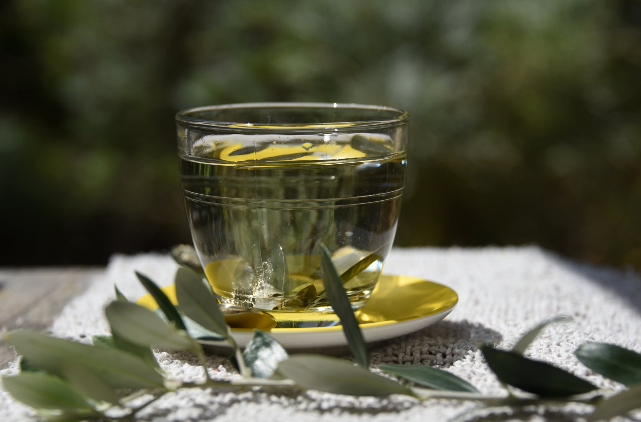 A clear glass cup and saucer containing olive leaf tea with an olive leaf laid on the table next to it.