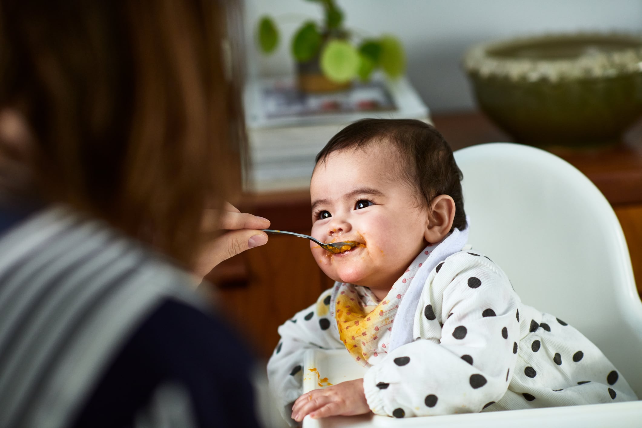 Baby girl in high chair eating baby food from spoon, dependency, care, enthusiasm