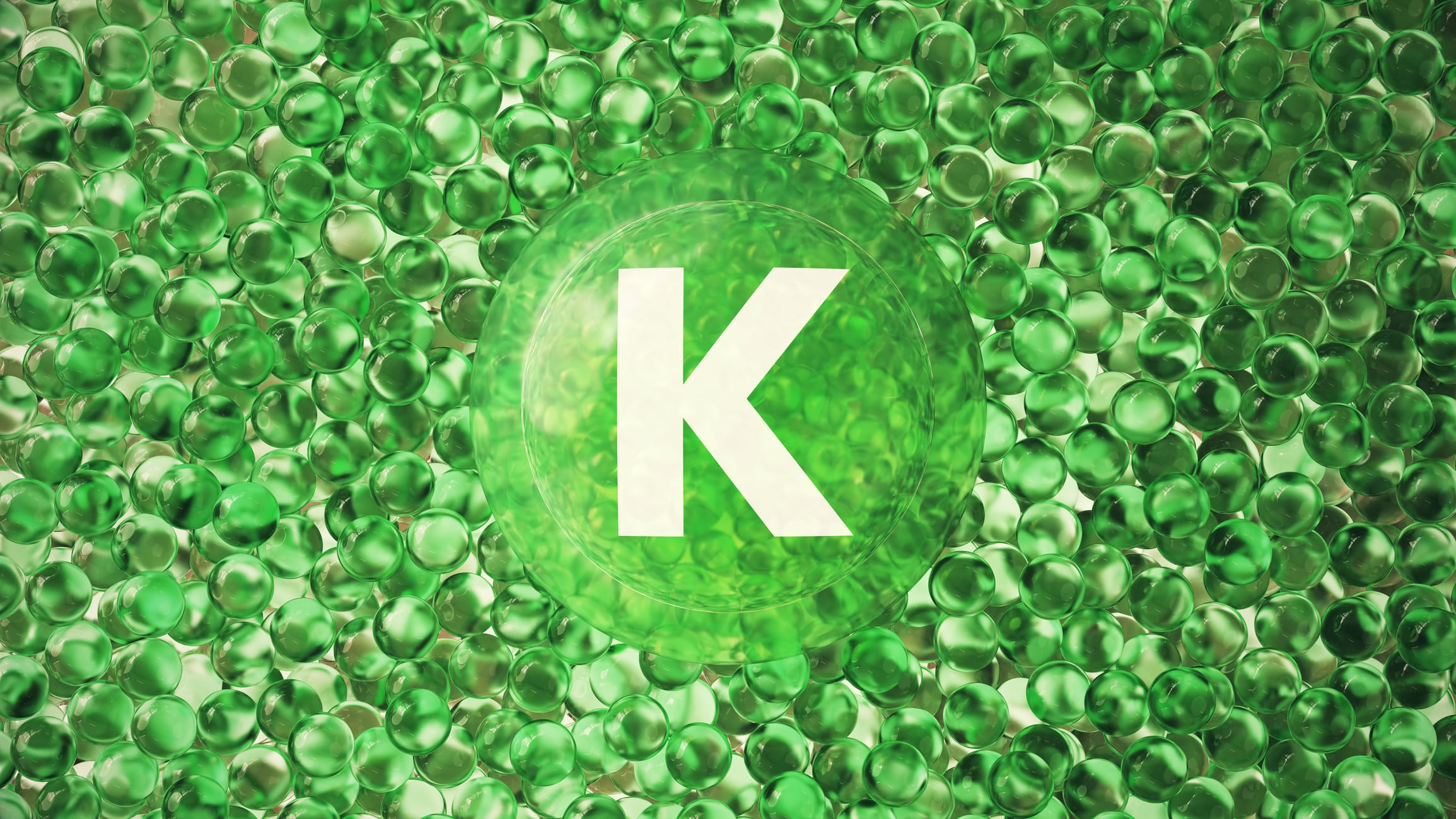 A modern 3D rendering featuring a large green transparent capsule marked with the letter "K", surrounded by small glossy gel-like spheres