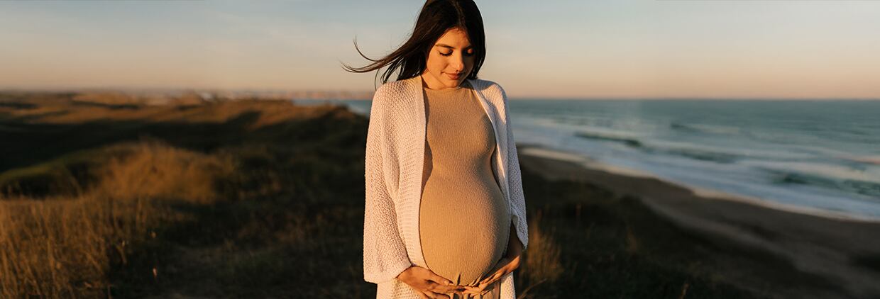 Crop serene pregnant female with long flying brown hair embracing belly while standing on grassy coast washed by sea waves at sunset
