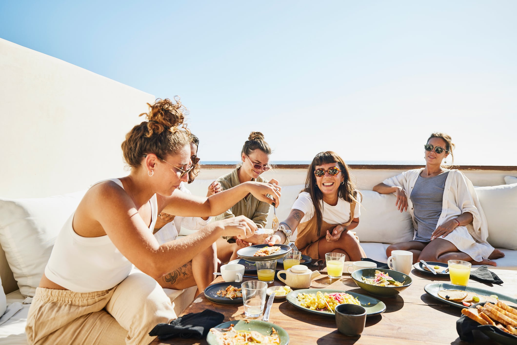 Medium wide shot of smiling female friends sharing breakfast on deck of luxury suite at tropical resort.