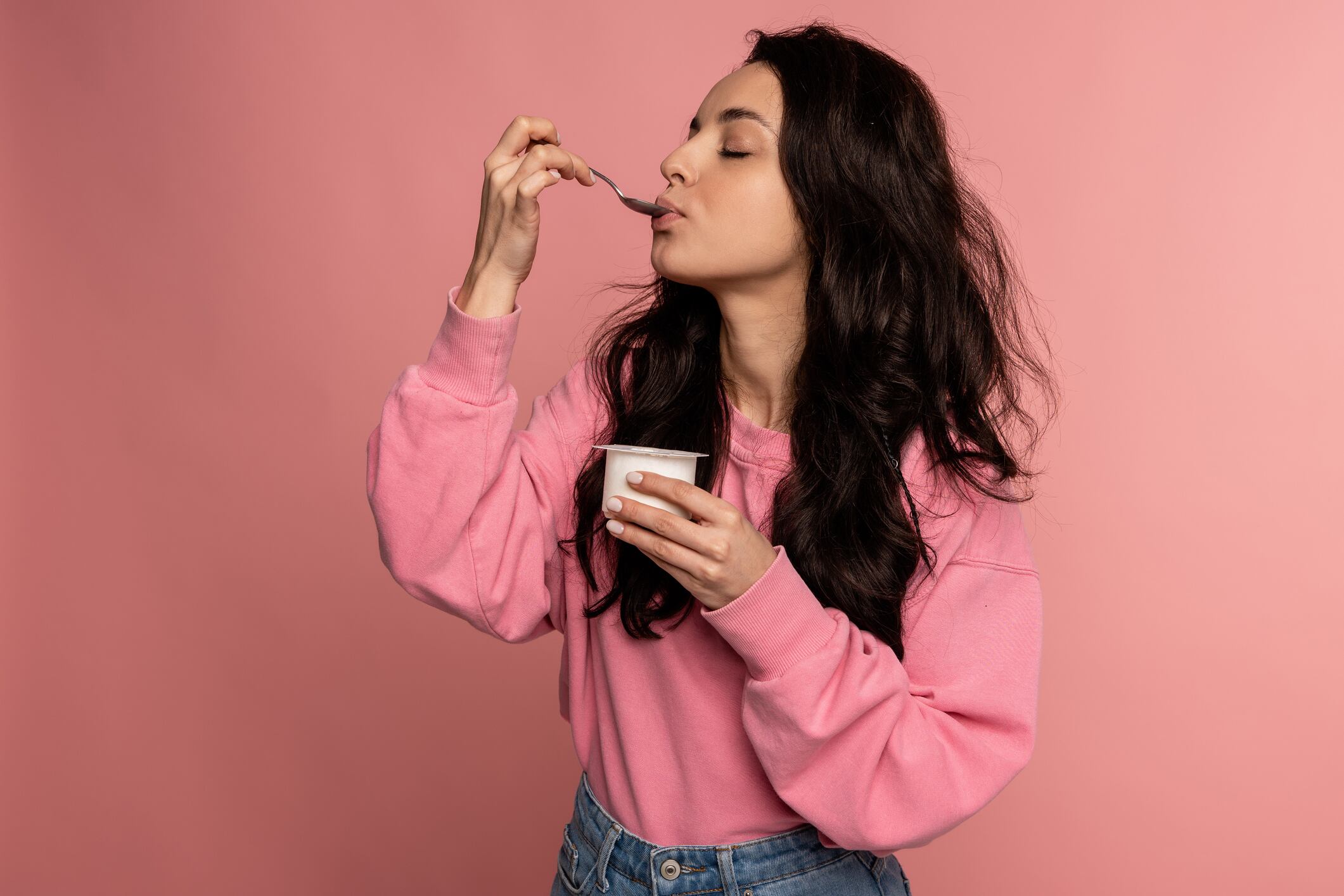 Waist-up portrait of a young woman snacking on the yogurt with a spoon from the plastic cup during the studio photo shoot. Healthy diet concept